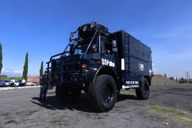 Polizeilaster auf einem Parkplatz neben einem Gebäude mit Menschen in der Nähe, Bäume und Gebäude im Hintergrund unter einem klaren blauen Himmel.