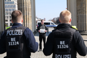 Drei Polizeibeamte stehen vor einem Gebäude mit einem Mann, der ein Papier zwischen ihnen hält, flankiert von Säulen, während Fahrzeuge, Menschen, Bäume und Gebäude den Hintergrund füllen.