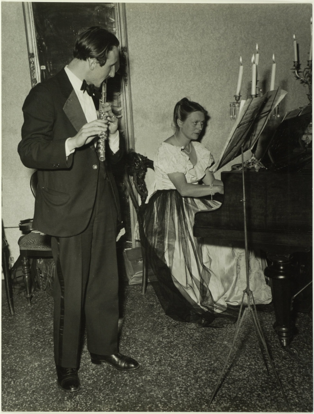 Mann im Smoking, der eine Klarinette spielt, neben einer Frau, die an einem Piano sitzt, mit Notenständer, gerahmtes Kunstwerk an der Wand im Hintergrund.