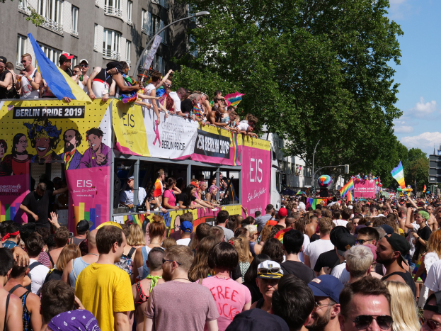 Eine große Menschenmenge marschiert auf der Straße während des Christopher Street Day 2019 in Berlin, viele tragen Mützen und Goggles, einige halten Fahnen, mit Bannern, Laternenmasten, Bäumen und Gebäuden im Hintergrund unter einem bewölkten Himmel.