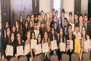 Gruppe von Menschen mit Urkunden bei einer Abschlussfeier, die auf Stühlen sitzen und eine Wand im Hintergrund haben.