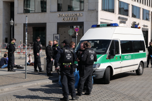 Polizisten stehen vor einem Starbucks-Café mit einem Van auf der rechten Seite und Menschen auf der linken Seite, während im Hintergrund ein Gebäude, ein Schild, ein Laternenpfahl und ein Zaun zu sehen sind.