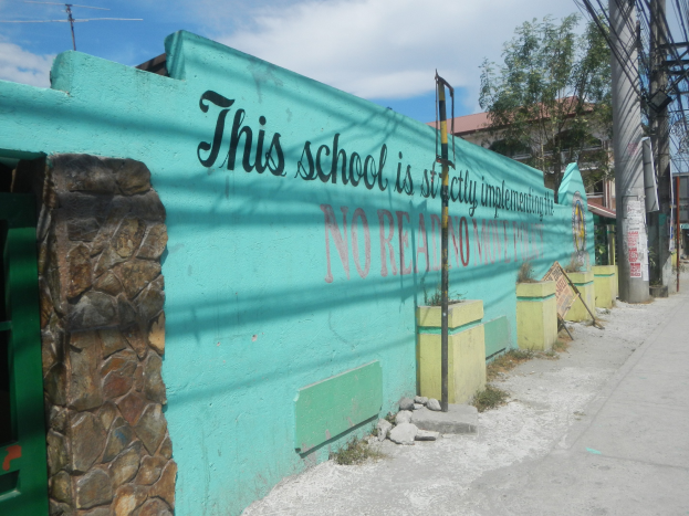 Grüne Wand mit Graffiti 'Diese Schule ist keine Stadt' umgeben von Gebäuden, Bäumen, Pfählen, Drähten und Brettern unter einem bewölkten Himmel.