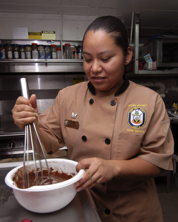 Eine Frau in Kochuniform steht in einer Küche und hält eine Schüssel mit einem Schneebesen in der Hand, umgeben von verschiedenen Gegenständen auf dem Tisch und Flaschen auf Regalen im Hintergrund.