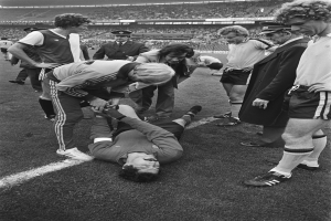 Schwarzes und weißes Bild eines Fußballspielers, der auf dem Boden liegt und von einer Gruppe von Männern umgeben ist, mit einer Menschenmenge im Hintergrund.