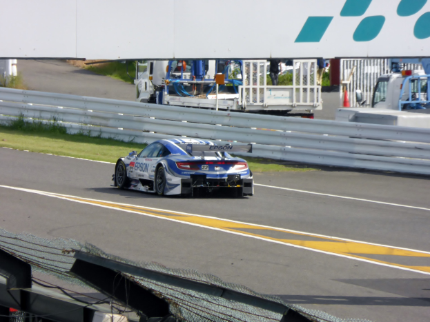 BMW M6 GT3 Rennauto auf einer Rennstrecke bei den 24 Stunden von Le Mans, umgeben von Absperrungen, Gras, Verkehrskegeln, Menschen und Bäumen unter einem klaren blauen Himmel.