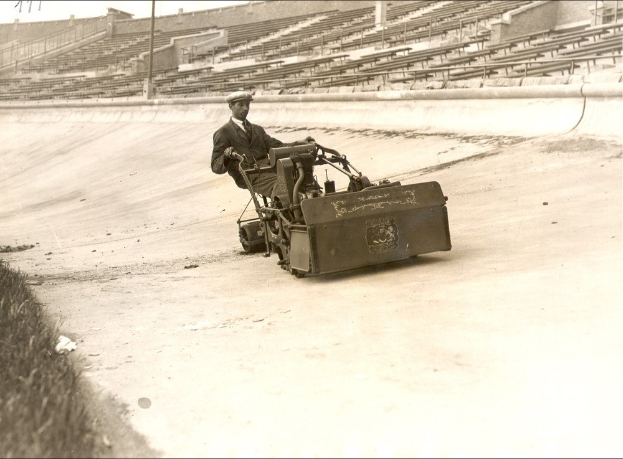 Altes Schwarz-Weiß-Foto eines Mannes, der einen Rasenmäher auf einer Straße mit Gras links und einem Stadion im Hintergrund fährt.