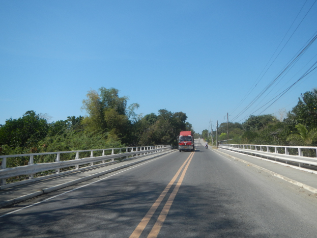 Ein roter Lkw fährt über eine Brücke mit Geländern, umgeben von Bäumen und Strommasten mit Drähten, unter einem klaren blauen Himmel.