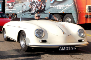 Ein Mann mit Schutzbrille fährt einen weißen Porsche 356 Speedster an einem roten Auto vorbei, wobei das Nummernschild deutlich zu sehen ist. Im Hintergrund befindet sich ein Lastwagen mit Bildern und Text.
