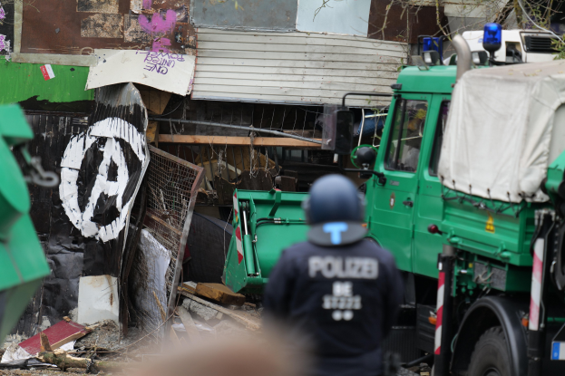 Ein Polizeibeamter steht vor einem mit Graffiti bedeckten Gebäude mit Fahrzeugen, Bäumen und anderen Gegenständen in der Nähe; ein Teil eines menschlichen Gesichts ist unten sichtbar.