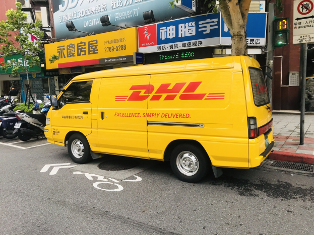 Gelber DHL-Lieferwagen auf der Straße geparkt, umgeben von Gebäuden, Bäumen, Schildern, Strommasten, Ampeln und einer Person auf dem Gehweg.
