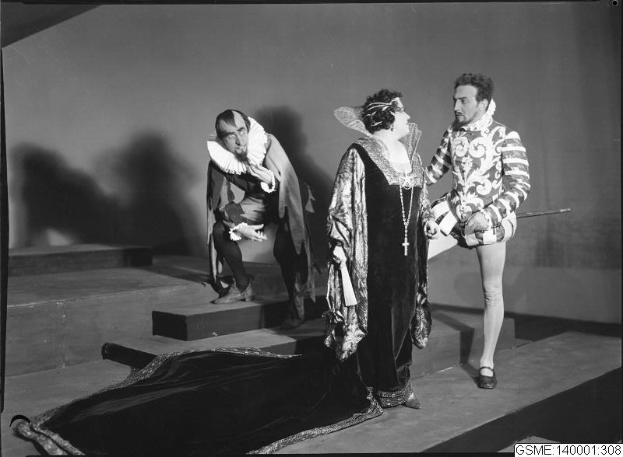 Schwarzes und weißes Foto von drei Menschen auf einer Bühne, wobei einer im Kostüm einen Stock hält, ein anderer auf einer Treppe sitzt und unten ein Text mit "Der Phantom der Oper" steht.