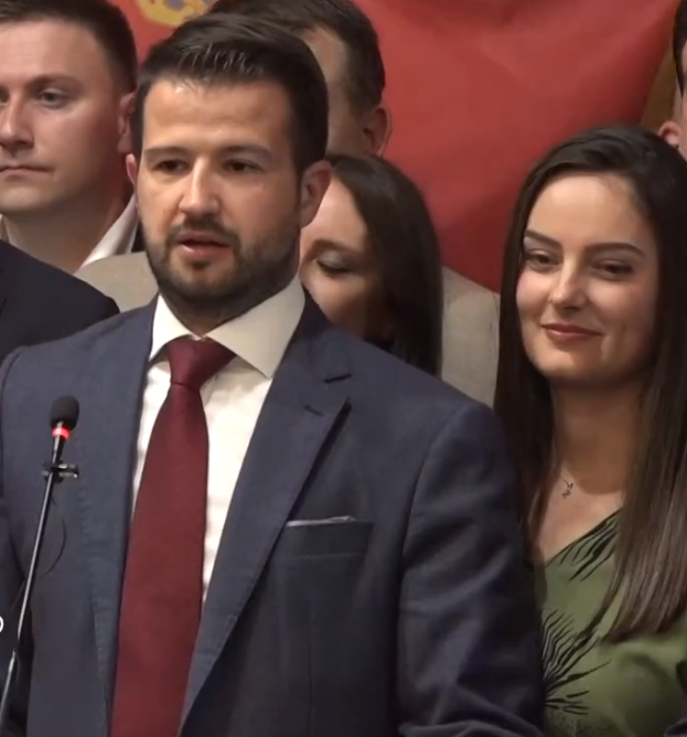 Ein Mann im Anzug und Krawatte steht an einem Podium mit einem Mikrofon und einer Fahne im Hintergrund.