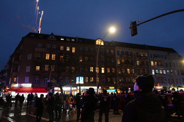 Eine Menschenmenge, die nachts eine von Straßenlaternen und Ampeln beleuchtete Straße entlanggeht, mit Gebäuden, Bäumen und Texttafeln im Hintergrund unter einem Feuerwerkhimmel.