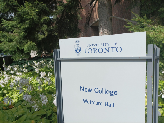 Außenansicht des neuen Colleges der University of Toronto an der Wetmore Hall, mit einem Schild vor dem Gebäude, umgeben von grünem Gras, bunten Blumen, hohen Bäumen und einem klaren blauen Himmel.
