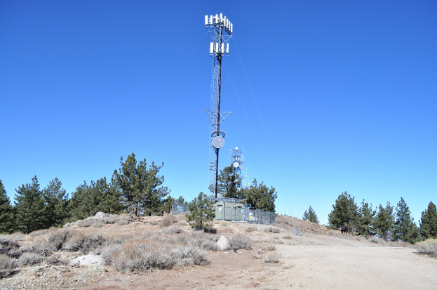 Ein Mobilfunkmast steht auf einem Schotterfeld mit Bäumen und Pflanzen im Hintergrund unter einem klaren blauen Himmel.