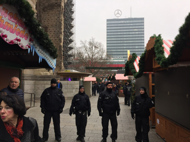 Eine Gruppe von Polizisten in schwarzen Uniformen und Mützen steht vor einem geschmückten Weihnachtsmarkt in Berlin, mit festlichen Dekorationen, Gebäuden, Bäumen und einem klaren blauen Himmel im Hintergrund; ein Schild mit Text ist links sichtbar.