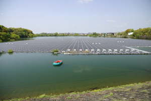 Kleines Boot mit Solarpanelen auf der Oberfläche, das auf einem Gewässer schwimmt und von Grünfläche umgeben ist, mit Gebäuden und einem klaren Himmel im Hintergrund.