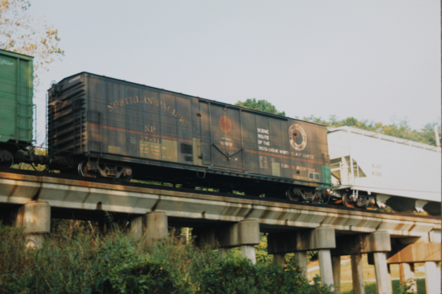 Ein G├╝terwagen ├╝berquert eine Br├╝cke, umgeben von saftigen Pflanzen und B├Ąumen, mit einem hellblauen Himmel im Hintergrund.