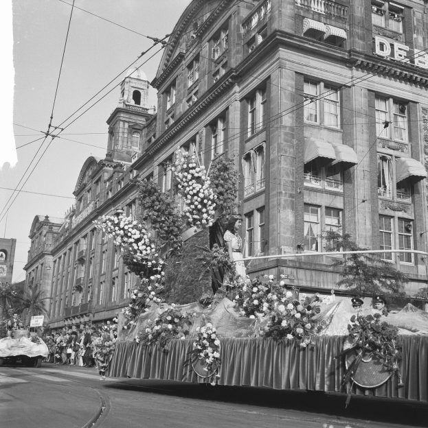 Ein Schwarz-Weiß-Foto eines Blumen-schmückenden Festwagen bei einem Umzug auf einer Stadtstraße, umgeben von Menschen, mit Gebäuden, Bäumen und einem klaren Himmel im Hintergrund.