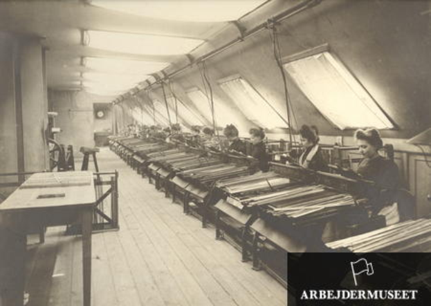 Ein Schwarz-Weiß-Foto von Frauen, die in einer Textilfabrik arbeiten, sitzend an Maschinen mit Tischen auf der linken Seite, eine Uhr an der Wand im Hintergrund und Text in der rechten unteren Ecke.