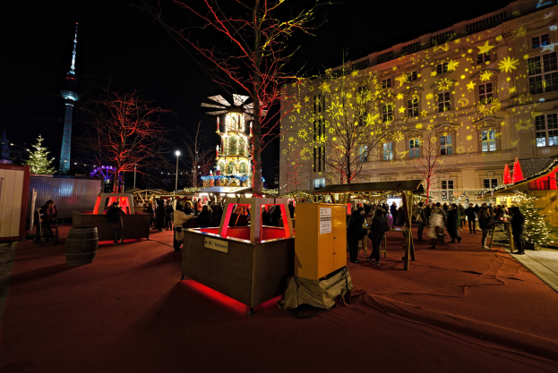 Ein belebter Weihnachtsmarkt in Berlin mit Menschen um geschmückte Stände, festliche Lichter, Bäume, Gebäude, Laternenmasten und einen Turm unter einem dunklen Himmel.