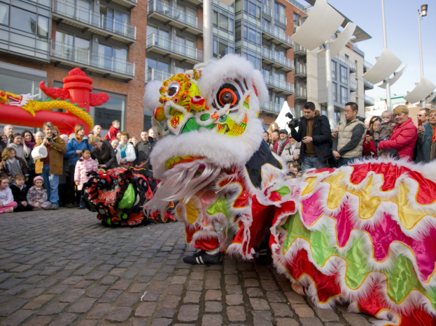 Ein lebhaftes chinesisches Neujahrsfest in Amsterdam mit einer Löwen-Tanz-Performance vor einer Zuschauermenge, darunter einige, die das Ereignis fotografieren, vor einem Gebäude und einem klaren blauen Himmel.