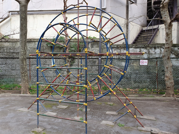 Spielplatz mit einem großen Metallklettergerüst, umgeben von Bäumen und einer Mauer mit Maschendrahtzaun, mit Treppen mit Geländern, die zu einem Gebäude mit Fenstern und Rohren führen, einem Schild an einem Pfahl und Grasboden.