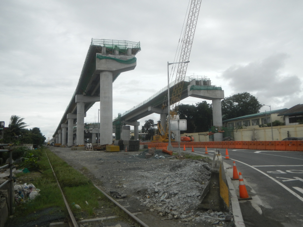 Baustelle mit Brücke im Hintergrund, Straße mit Verkehrskegeln markiert, verstreute Steine und Gras, eine Eisenbahnschiene links, Bäume und Gebäude säumen die Straße und ein bewölkter Himmel.