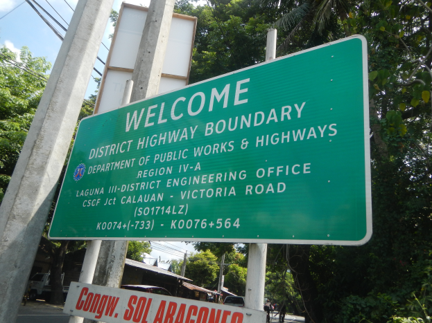 Ein grünes Schild mit der Aufschrift 'Willkommen an der Kreisgrenze' ist an zwei weißen Pfählen befestigt, im Hintergrund sind Fahrzeuge, Gebäude und Bäume unter einem blauen und weißen Himmel zu sehen.