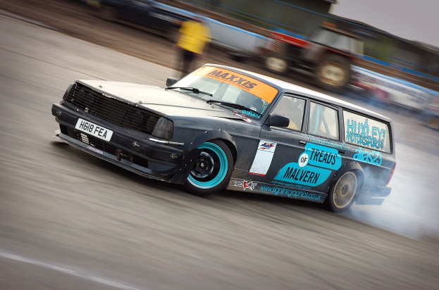 Ein Auto beim Rennsport auf einer Rennstrecke, das Rauch ausstößt, mit anderen Fahrzeugen und Zuschauern im Hintergrund.