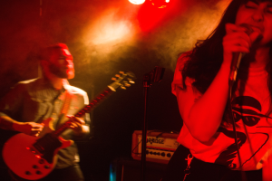 Eine Frau singt in ein Mikrofon auf der Bühne, während ein Mann im Hintergrund Gitarre spielt und Deckenlampen die lebendige Kulisse beleuchten.