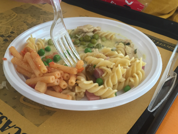 Eine Person, die eine Gabel über eine Schüssel mit Nudeln, Erbsen und Schinken hält, mit einem Tablett mit einem Löffel und einer Abdeckung auf dem Tisch.