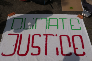 Weißer Banner mit dem Text "Klimagerechtigkeit" auf dem Boden vor Pappe, einer Tüte und anderen Gegenständen.