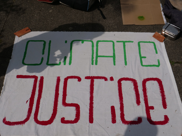 Weißer Banner mit dem Text "Klimagerechtigkeit" auf dem Boden vor Pappe, einer Tüte und anderen Gegenständen.