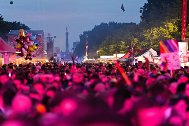 Eine Große Menge Menschen Auf Einem Nachtsmusikfestival, Beleuchtet Von Farbigen Lichtern, Mit Zelten, Fahnen Und Masten Im Vordergrund Und Bäumen, Einer Turm Und Einem Klaren Himmel Im Hintergrund.