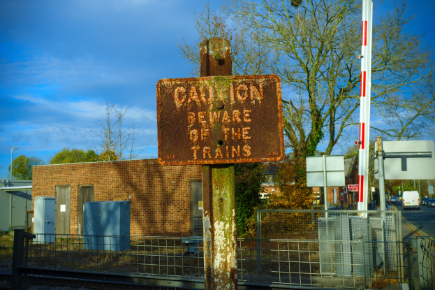 Achtungsschild auf einem Bahngleis, umgeben von einem Metallzaun, Bäumen, Strommasten, einem Gebäude mit Fenstern, Containern, einem Straßenmast, Fahrzeugen auf einer Straße und einer bewölkten Himmel.