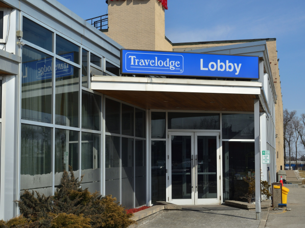 Außenansicht des Travelodge Hotels in Ottawa, Ontario, mit Glaswänden und -türen, einem Schild, Topfpflanzen vorne und Bäumen vor einem klaren blauen Himmel.