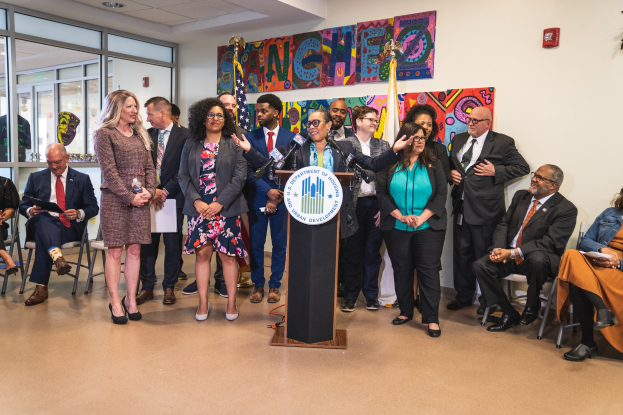 Gruppe von Menschen vor einem Podium mit einem Mikrofon und einer Tafel mit der Aufschrift "Sancho Stadtrat tritt dem Stadtrat bei", mit sitzenden und stehenden Teilnehmern, Flaggen, Feuerlöscher und Deckenbeleuchtung im Hintergrund.