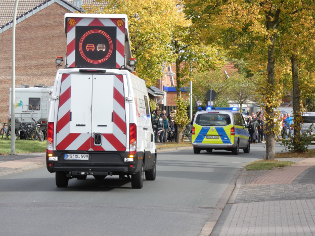 Ein Rettungswagen fährt eine von Bäumen gesäumte Straße mit Menschen auf dem Bürgersteig, Fahrrädern, Pfählen und Gras.