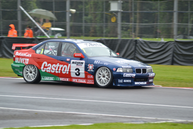 Ein Auto auf einer Rennstrecke mit Text und Zahlen, umgeben von Gras, mit Maschendrahtzaun und schwarzem Tuch im Hintergrund, Menschen in orangefarbenen Jacken mit Schirmen und Bäumen.