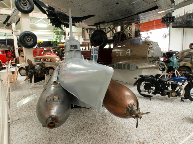 Großes Flugzeug in einem Museum ausgestellt, umgeben von verschiedenen Fahrzeugen und einer Begrenzung auf der linken Seite sowie Bäumen und Deckenleuchten im Hintergrund.