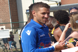 Ein Mann in einem blauen Hemd signiert Autogramme für eine Gruppe von Menschen, einige tragen Kappen und Schutzbrillen, vor einem Gebäude mit Fenstern, Maschendrahtzaun und einem Fahrrad.