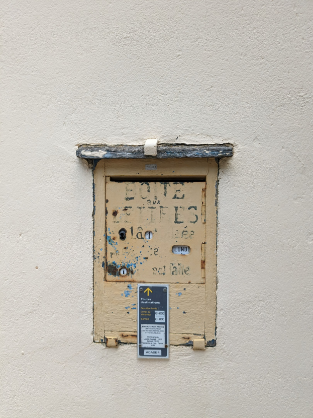 Alte rechteckige Metall-Türklingel mit Schlüsselloch und beschrifteter Vorderseite, an einer verwitterten Gebäudewand mit sichtbaren Rissen und Absplitterungen montiert.