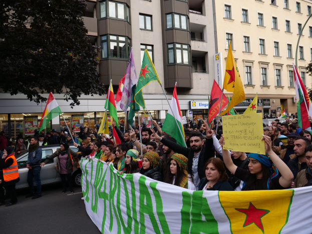 Große Gruppe von Menschen marschiert auf einer Straße mit Fahnen und Spruchbändern, mit einem geparkten Auto auf der rechten Seite und einem Baum auf der linken Seite, vor Gebäuden mit sichtbaren Fenstern und Schildern.