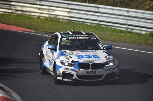 Ein BMW M4 DTM-Rennwagen mit Text und Zahlen auf der Karosserie fährt auf einer von Gras, einem Zaun und Pflanzen umgebenen Strecke, wobei ein Rad sichtbar ist.