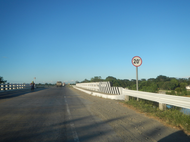 Fahrbahn mit Fahrzeugen, Geschwindigkeitsbegrenzungsschild, Geländer, Gras, Bäume und klarer blauer Himmel.