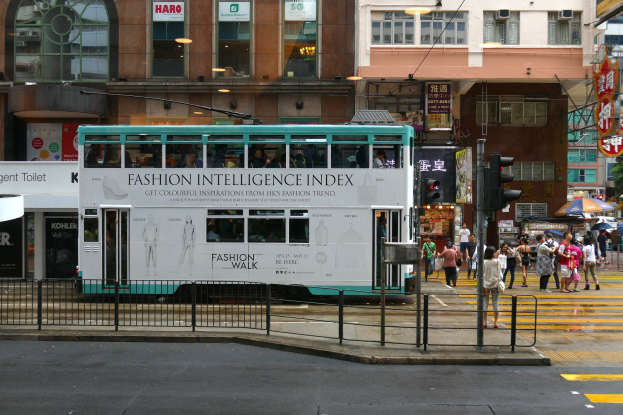Ein Doppeldeckerbus fährt eine Stadtstraße mit hohen Gebäuden, Fußgängern auf dem Gehweg mit Schirmen, Verkehrsampeln, einem Gehweggeländer und Texttafeln sowie Bäumen und einem klaren blauen Himmel im Hintergrund.