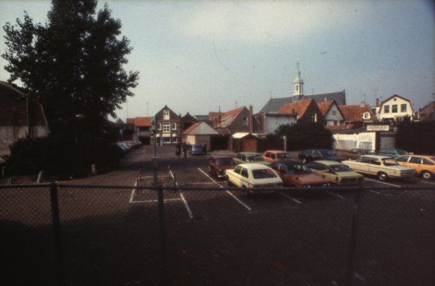Altes Schwarz-Weiß-Foto eines umzäunten Parkplatzes mit parkenden Autos, Bäumen, Strommasten, umgebenen Gebäuden, einigen Menschen und Himmel im Hintergrund.