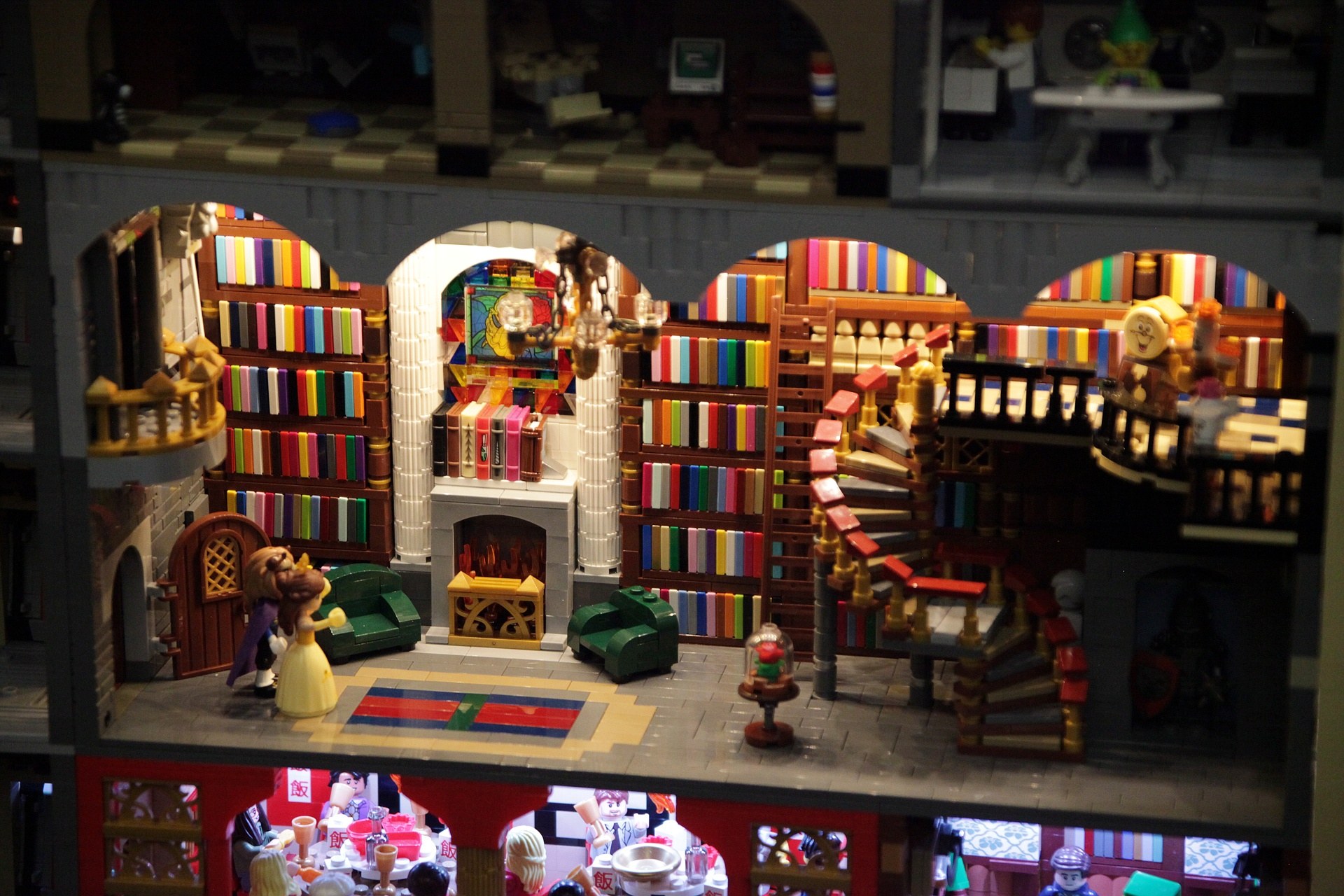 Lego-Modell einer Bibliothek mit mehreren Bücherregalen, einer Treppe, einem Kronleuchter, einem Kamin, einer Sitzgruppe, einem Tisch mit Stühlen, einem Teppichboden und verstreuten Spielzeugen.